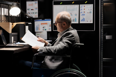 Professional accountant in wheelchair checking administrative paperwork in dark file room. Businessman in bureaucratic file cabinet office filled with invoice folders and flowchartsの写真素材