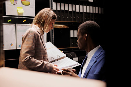 Diverse depository employees analyzing bureaucracy record, checking administrative documents late at night in storage room. Bookkeepers discussing accountancy report, discovering management filesの写真素材