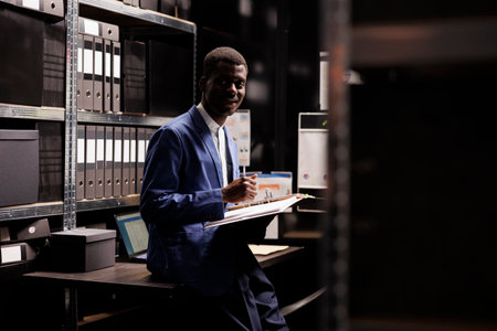 Private detective working at confidential case, analyzing files with evidence about crime scene in arhive room. African american police officer checking criminology report late at nightの写真素材