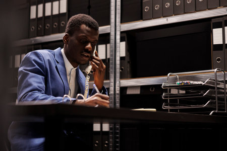 African american police officer explaining criminology report to remote investigator officer using landline phone. Private detective working overhours at crime case, trying to catch mysterious suspectの写真素材