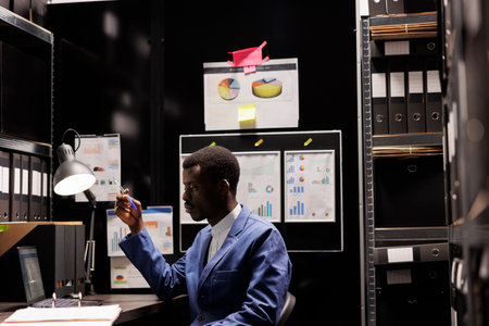 African american police officer analyzing criminology report, checking crime scene files on laptop computer. Private investigator working overtime in arhive room, looking at investigation evidenceの写真素材