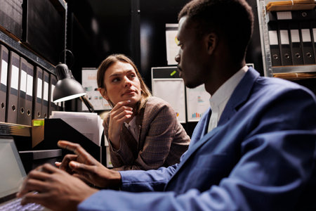 Investigation team working overhours at fraude case, analyzing new evidence in arhive room. Diverse private detectives reading police expertise, trying to catch suspect. Criminology conceptの写真素材