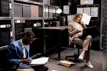 Tired diverse inspectors working overhours in police station, analyzing criminal case evidence. African american private detectives checking crime scene footage trying to catch mysterious suspectの写真素材