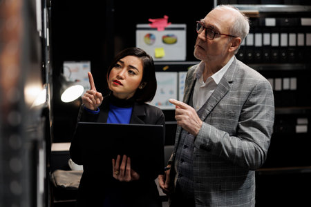 Teamworking professional accounting elderly businessman and asian coworker looking for bookkeeping bureaucratic paperwork on file cabinet shelves. Team executives in storage repository office roomの写真素材