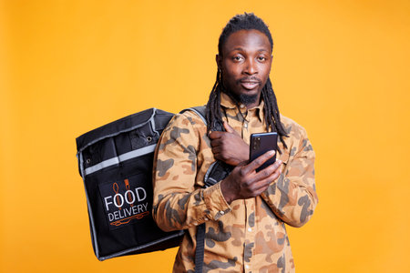 Pizzeria courier checking customers adreess on phone using takeaway food app before delivery food order. African american restaurant employee carrying thermal backpack standing in studioの写真素材