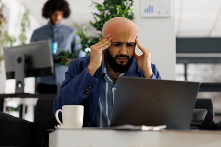 Start up product manager with headache working to meet deadlines in business office. Arab company entrepreneur suffering from migraine and rubbing temples at workplace in coworking spaceの写真素材