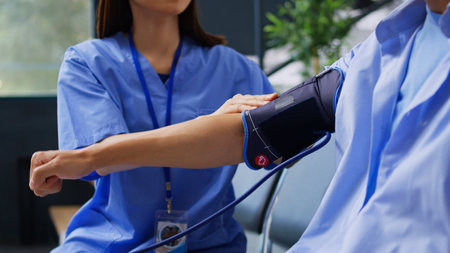 Assistant doing hypertension examination with tonometer in hospital waiting room, measuring asian...