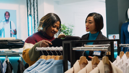 Asian employee showing clothes to customer in retail shop and buying formal wear, talking about new fashion collection. Store assistant working in shopping center, client shopper. Handheld shot.の写真素材