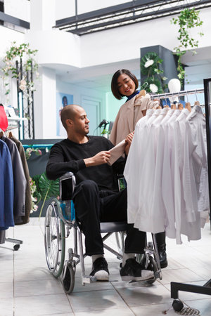 Clothing store seller offering help to arab man client with physical impairment in choosing formal jacket for woman. Boutique employee showing trendy garment to customer in wheelchairの写真素材