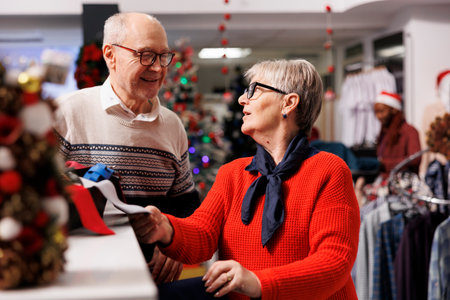 Cheerful elderly couple shopping at mall, looking to buy formal attire for christmas eve dinner outfit. Senior people checking ties from accessories box at clothing store, xmas decorations.の写真素材