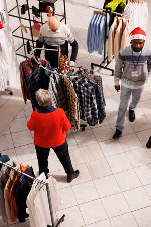 Various shoppers looking at clothing items on racks in shopping center while seeking for garments to purchase. Consumers going apparel shopping as gifts for festive season, Christmas eve.の写真素材