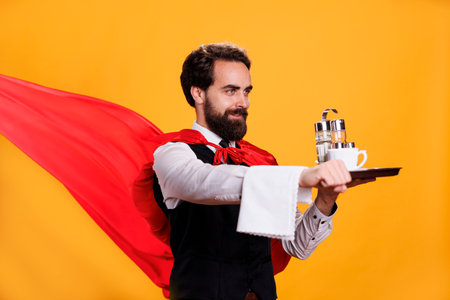 Superhero waiter carries food tray with red cape fluttering on camera, young man working at restaurant as butler serving dinner against yellow background. Professional elegant server.の写真素材