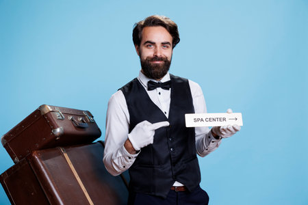Smiling bellhop poses with spa sign to guide visitors and present different hotel areas, tourism industry. Cheerful professional doorman holding indicator as direction for relaxation area.の写真素材