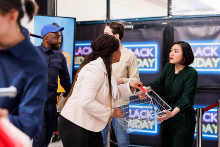 People fight for buying things during Black Friday. Two greedy angry diverse women shoppers standing at shopping mall entrance fighting for last shopping basket while hunting for bargainsの写真素材