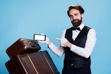 Bellboy presents blank screen for tourism industry against blue background. Hotel concierge in formal attire and gloves holds telephone with white copyspace display, posing on camera.の写真素材