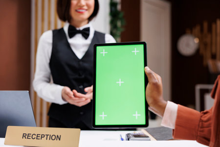 Adult with greenscreen at front desk, talking to reception staff about hotel room service and holding device. Guest looking at chroma key display with isolated mockup template in lobby.の写真素材