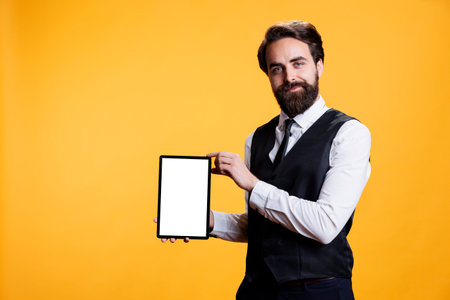 Young waiter holding tablet with white screen on camera, presenting isolated empty template against yellow background. Professional employee shows device with blank mockup display.の写真素材