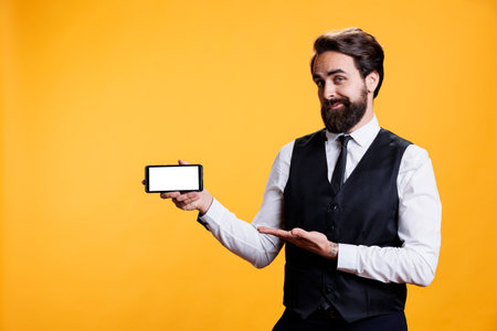 Young butler shows white display on camera, holding smartphone with isolated copyspace screen. Professional skilled restaurant employee presents blank empty screen template in studio.の写真素材