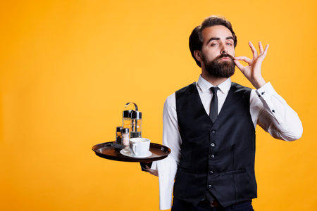 Private young man does sealed lips sign or hush mute gesture in studio, posing on yellow background. Professional restaurant worker presenting silence gesture, trying to keep secret.の写真素材