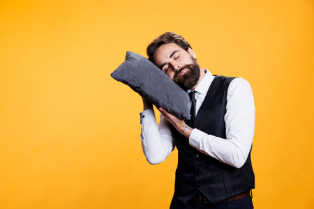 Sleepy butler sleeping on pillow, taking a nap while he in standing in studio. Young exhausted overworked person falling asleep against yellow background, working in restaurant industry.の写真素材