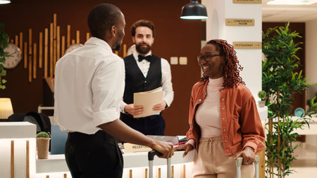 African american couple checking out from luxury hotel after successful honeymoon vacation. Happy guests assisted by friendly helpful front desk receptionist in resort lobbyの写真素材