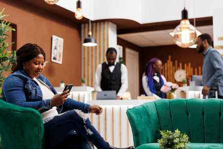 Happy relaxed African American woman relaxing in hotel lobby using smartphone watching online video or browsing internet. Female tourist resting with phone in lounge area at luxury resortの写真素材
