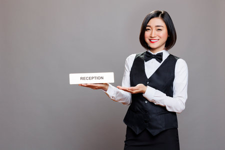 Restaurant receptionist showcasing reception steel plate, smiling and looking at camera with happy expression. Hotel asian woman worker showing tabletop sign studio portraitの写真素材
