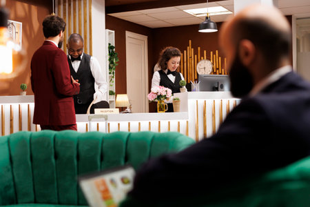 Front desk staff greeting businessman with room reservation, talking about services in lobby. White...