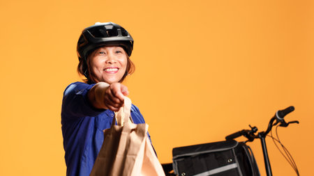 Courier waiting in front of house door for client to pick up takeout meal order, close up. Asian delivery woman offering takeaway food bag to customer, isolated over orange studio backgroundの写真素材