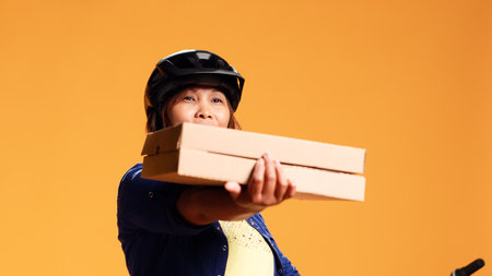 Hip level close up shot of cheerful courier looking at camera, showing approval gesture while delivering takeout meals. Friendly cyclist offering pizza to customer, isolated over studio backgroundの写真素材
