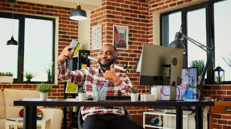 Happy cheerful african american man taking selfie while smiling and showing thumbs up sign. Man in nice stylish modern house capturing photo using smartphone while remotely working from homeの写真素材