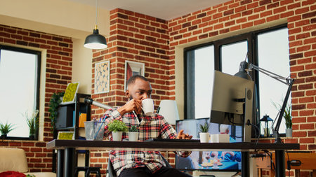 Relaxed cheerful remote worker enjoying cup of coffee while working from home. Smiling freelancer typing on computer, listening to TV show in stylish cozy apartment living roomの写真素材