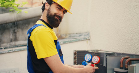 Efficient technician looking at refrigerant levels in external air conditioner while using manifold meters to precisely measure the pressure in HVAC system, ensuring efficient cooling performanceの写真素材