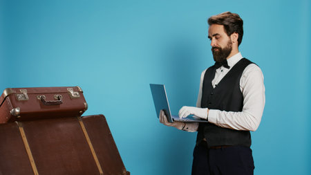 Hotel staff reviews list of guests on online bookings while using laptop and standing next to stack of suitcases. Employee working as doorkeeper providing an enjoyable interaction.の写真素材