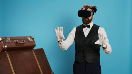 Modern doorman using vr glasses against blue background, using virtual reality technology on headset in studio. Young bellboy with classy occupation standing next to trolley bags, travel sector.の写真素材