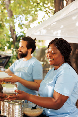 Close up image of african american female volunteer handing out canned goods to homeless people in need. Black woman and caucasian man share donation boxes filled with canned goods at food drive.の写真素材