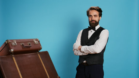 Hotel porter with baggage in studio, providing outstanding service to carry luggage for guests. Professional bellhop or doorman with suit and tie feeling confident and determined on camera.の写真素材