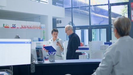 Elderly man in need of prescription medicaments informations in apothecary, requesting assistance from cashier who redirects him to qualified chemist checking pharmaceutical products inventoryの写真素材