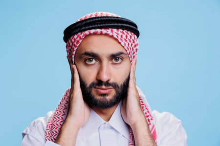 Muslim man closing ears with palms while showing hear no evil three wise monkeys concept studio portrait. Arab person wearing traditional clothes showcasing hearing problems and looking at cameraの写真素材