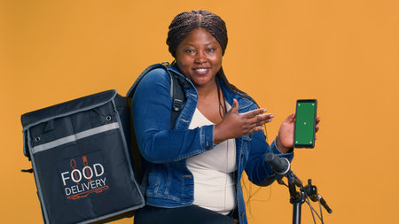 Lady with african american ethnicity showing cell phone with blank chromakey mockup template. Technology-driven delivery person grasps mobile phone with her left-hand displaying green screen.の写真素材