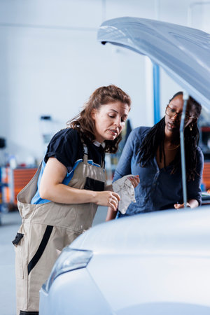 Engineer in garage finishing fixing car for BIPOC woman, looking underneath vehicle hood to remove remaining oil leaks. Worker does routine exhaust system cleaning on client automobileの写真素材