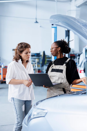 Organized mechanic next to client in car service orders new engine for damaged vehicle using laptop. Employee looking for components on internet to replace old ones in faulty automobileの写真素材