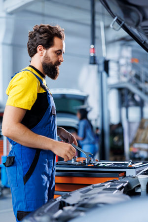 Serviceman in car service picks wrench from work station bench, using it to unscrew nuts in order to replace engine. Precise garage worker uses professional tools to fix customer automobileの写真素材