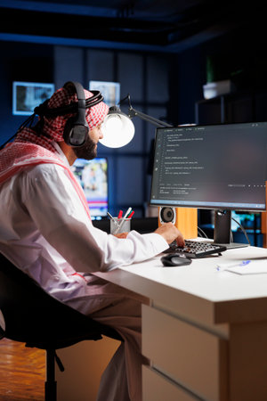 Data developer from the Middle East types code on a computer, demonstrating the core of software programming and development. Muslim man using wireless headphones and immersed in algorithm execution.の写真素材