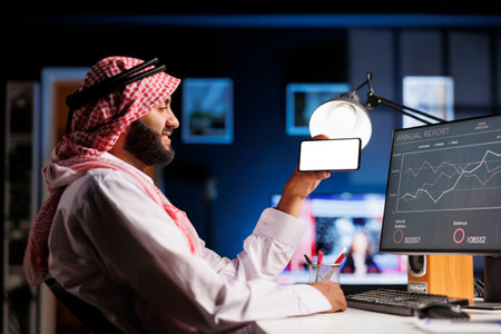 An Arabic individual, wearing traditional clothing uses a mobile phone at an office desk. The isolated white screen offers space for customized content in an advertising or promotional setting.の写真素材