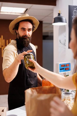 Woman at checkout counter in zero waste shop, buying locally sourced ecofriendly vegetables from happy storekeeper. Client purchasing pickles jar in local neighborhood store using renewable energyの写真素材