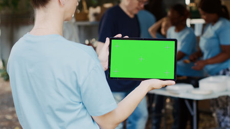 Caucasian woman horizontally holding digital tablet with green screen chromakey display at an outdoor food bank. Smart device displaying blank mockup template held by female charity worker, outdoors.の写真素材