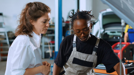 African american engineer assisting customer with car maintenance in repair shop. Trained professional in garage looking over car components with woman, servicing her vehicle during annual checkupの写真素材
