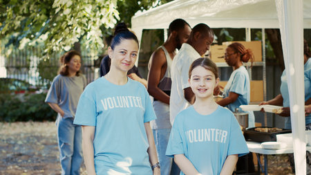 Portrait shot of mother and daughter participating in hunger relief program at outdoor food bank. Caucasian volunteers in blue t-shirts and looking at camera, ready to assist the needy. Tripod shot.の写真素材