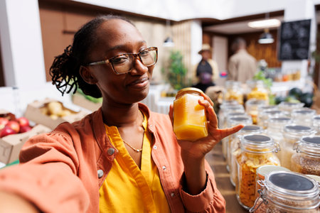 During video call, african american lady with glasses clutches a glass jar of honey. At eco friendly store, black woman uses phone for vlogging and leverages technology for internet marketing.の写真素材
